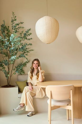Een vrouw in een beige pak zit glimlachend aan een houten tafel in een moderne, minimalistische kamer met lichte muren, papieren hanglampen die aan het plafond hangen en een Kunstplant Eucalyptus van 180 cm die de ruimte wat groen geeft.