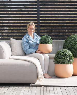 Een vrouw zit op blote voeten op een lichtgekleurde buitenbank, glimlacht en houdt een Kunstplant Buxus romeinse stijl UV D30cm vast. Twee soortgelijke kunstplanten staan vlakbij, met houten lattenhekwerk en terrasdecor op de achtergrond.