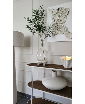 Een minimalistische consoletafel toont een heldere vaas met de Kunst Olijftak 109 cm, een ronde vaas van wit keramiek en een witte schaal met LED-kaarsen. Erboven hangt gestructureerde witte wandkunst en vlakbij staat een staande lamp.