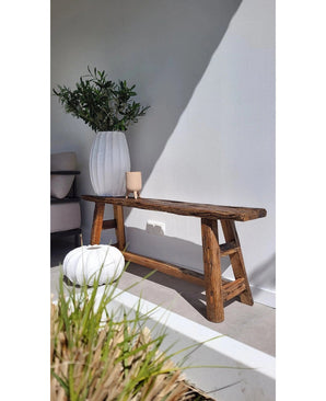 Een rustieke houten bank staat tegen een witte muur, naast een hoge witte vaas met Kunst olijfblad 90 cm UV voor een mediterrane sfeer. Vlakbij staan een ronde witte poef en een kleine kandelaar, met zonlicht dat zachte schaduwen werpt.