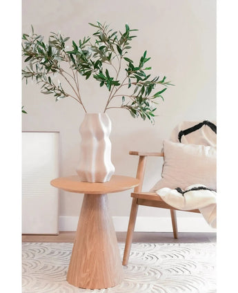 Ein modernes Wohnzimmer mit einem Holzstuhl mit hellem Kissen, einem runden Beistelltisch und einer weißen skulpturalen Vase mit künstlichem Olivenblatt 90 cm UV, das einen minimalistischen Raum mit sanften Neutraltönen und einer subtilen mediterranen Atmosphäre schafft.