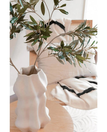 Auf einem hellen Holztisch steht eine weiße, skulpturale Vase mit künstlichem Olivenblatt 90 cm feuerfest. Dahinter sitzen zwei cremefarbene Kissen mit schwarzen Streifen auf einem beigen Stuhl und verleihen diesem modernen Raum gemütlichen Komfort.