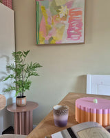 In einem pastellfarbenen Raum gibt es eine 85 cm große künstliche Farnpflanze auf einem rosa Beistelltisch, eine lila-goldene Tasse, eine rosa Schale mit Wellenrand, ein offenes Notizbuch und an der Wand ein abstraktes Gemälde mit Grün-, Rosa- und Gelbtönen.