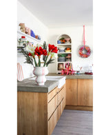 Een lichte keuken met houten kastjes, open witte planken met borden en potten, een feestelijke krans aan de muur en een Kunstbloem Amaryllis Real Touch rood 36 cm op het betonnen aanrecht.