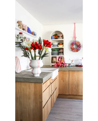 Een lichte keuken met houten kastjes, open witte planken met borden en potten, een feestelijke krans aan de muur en een Kunstbloem Amaryllis Real Touch rood 36 cm op het betonnen aanrecht.
