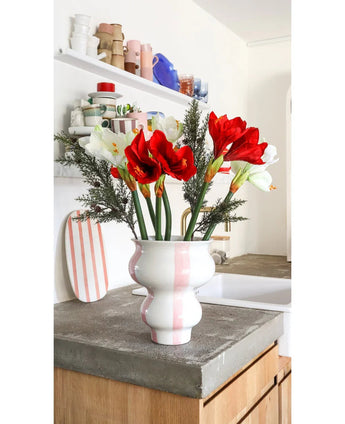 De Kunstbloem Amaryllis Real Touch rood 36 cm staat in een wit met roze gestreepte vaas van keramiek op een betonnen aanrecht in een lichte keuken met planken met kleurrijk servies op de achtergrond.