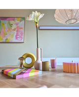 Een moderne houten tafel toont kleurrijke objecten, waaronder een roze vaas met daarin de Kunstbloem Amaryllis Real Touch wit 66 cm, een gele ringsculptuur, gestreepte boeken en een roze beker. Abstracte kunst accentueert de achtergrondmuur.