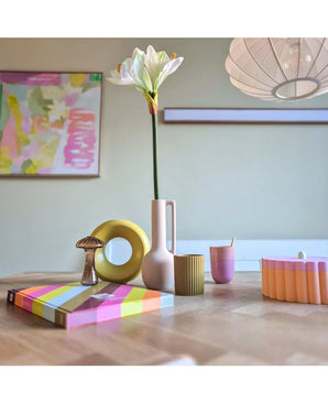 Een moderne houten tafel toont kleurrijke objecten, waaronder een roze vaas met daarin de Kunstbloem Amaryllis Real Touch wit 66 cm, een gele ringsculptuur, gestreepte boeken en een roze beker. Abstracte kunst accentueert de achtergrondmuur.