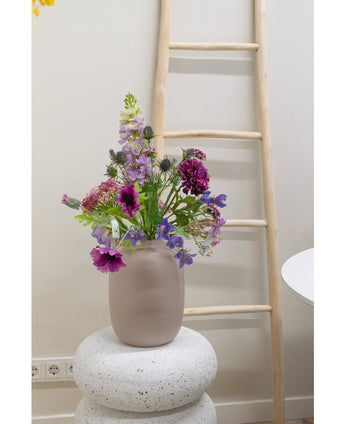 Een Kunstbloem Grasklokje van 62 cm paars staat in een matglazen vaas op een gestructureerde witte sokkel. Daarachter leunt een lichte houten ladder tegen een beige muur, met stopcontacten zichtbaar bij de vloer.