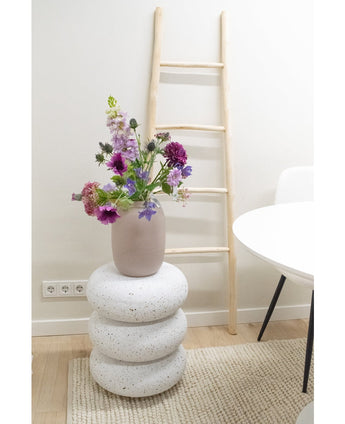 Een Kunstbloem Grasklokje van 62 cm paars staat in een vaas op drie ronde, witte steenachtige kussens. Daarachter staat een lichthouten ladder tegen een witte muur, naast een witte tafel en stoel op een beige vloerkleed.