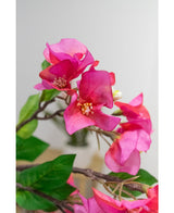 Close-up van de Kunstbloem Bougainvillea 90 cm paars: levendige paarse kunst bougainvillea bloemen met groene bladeren op een tak, tegen een zachte, lichte achtergrond.