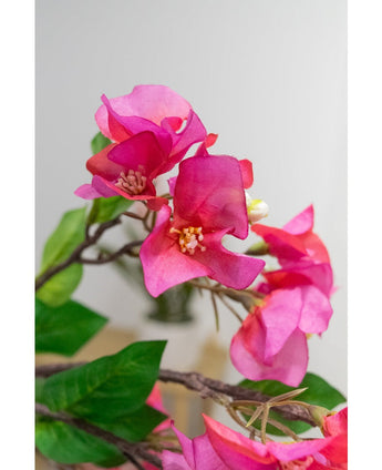 Close-up van de Kunstbloem Bougainvillea 90 cm paars: levendige paarse kunst bougainvillea bloemen met groene bladeren op een tak, tegen een zachte, lichte achtergrond.