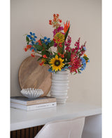 Een Kunstbloem Anjer tros 70 cm blauw in een vaas van wit keramiek staat op een wit bureau naast gestapelde boeken en een kleine decoratieve schaal, met daarachter een rond houten dienblad dat tegen de muur leunt.