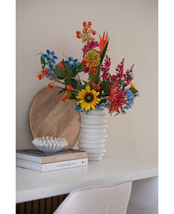 Ein künstlicher Nelkenstrauß 70 cm blau in einer weißen Keramikvase steht auf einem weißen Schreibtisch neben gestapelten Büchern und einer kleinen Dekoschale, dahinter lehnt ein rundes Holztablett an der Wand.
