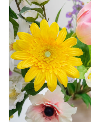 De Kunstbloem Gerbera 53 cm geel, een levendige gele kunstbloem met gelaagde bloemblaadjes, staat tussen andere kleurrijke kunstbloemen en groene bladeren, met witte en roze bloemen op de achtergrond.