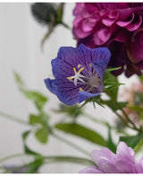 Een close-up van het Kunstbloem Grasklokje 62 cm paars, een delicate paarse kunstbloem met diepe nerven en gele meeldraden, tussen groene bladeren en zacht vage roze en paarse bloemen op de achtergrond.