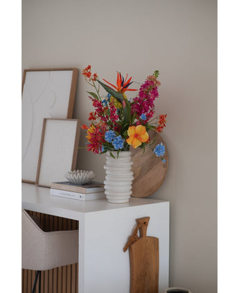 Eine weiße gerippte Vase mit künstlicher Katzenminze in Rosa (75 cm) steht auf einem weißen Schreibtisch mit Büchern, gerahmter Kunst, einem Holztablett und einem Schneidebrett neben einem beigen Stuhl in einem modernen, minimalistischen Raum.