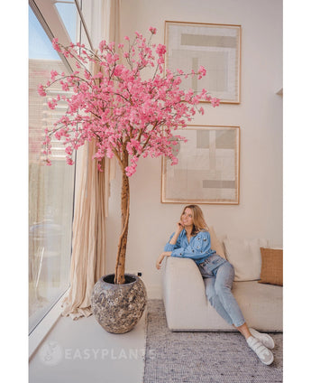 Een vrouw in casual kleding ontspant op een beige bank naast een 240 cm Kunstboom Kersenbloesem roze met roze bloemen, geplaatst in een moderne woonkamer met abstracte muurkunst en grote ramen.