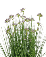 De Kunstgras Prei Bloem Ui Graspot 60cm heeft hoge, dunne groene bladeren en trossen kleine, delicate lichtpaarse bloemen op lange stelen. Het arrangement is brandvertragend en afgebeeld tegen een witte achtergrond.