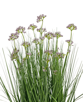 De Kunstgras Prei Bloem Ui Graspot 60cm heeft hoge, dunne groene bladeren en trossen kleine, delicate lichtpaarse bloemen op lange stelen. Het arrangement is brandvertragend en afgebeeld tegen een witte achtergrond.