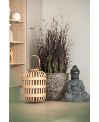 Auf einer hellen Fläche, in der Nähe eines Holzregals und einer hellen Wand, sind eine Holzlaterne, ein hoher Steintopf mit 100 cm künstlichem Zwiebelgras-Bonsai (feuerhemmend) und eine graue Buddha-Statue angeordnet.