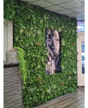Een wand bekleed met Kunsthaag mat Savanne 100x100 cm, met UV-beschermd groen in een kunststof plantenbak Ø100, heeft een groot zwart-wit portret in het midden. Het moderne interieur heeft een betegeld plafond en een houten balie aan de linkerkant.