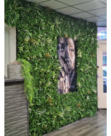 Een wand bekleed met Kunsthaag mat Savanne 100x100 cm, met UV-beschermd groen in een kunststof plantenbak Ø100, heeft een groot zwart-wit portret in het midden. Het moderne interieur heeft een betegeld plafond en een houten balie aan de linkerkant.