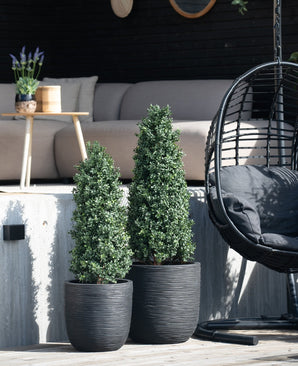 Twee Kunstplant Buxus Pyramide 120 cm UV-struiken staan op het terras. Een beige buitenbank, een ronde houten tafel met een plant en een zwarte hangende eierstoel erachter zorgen voor UV-bescherming en creëren een moderne, gezellige tuinscène.