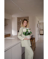 Een vrouw met krullend blond haar, gekleed in een lichtgroene outfit, staat in een moderne keuken met een Kunstplant Hortensia Wit UV 35 cm in haar handen – met crèmewitte bloemen – en glimlacht naar de camera.