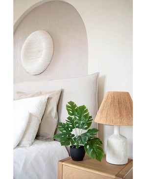 Een moderne slaapkamer in neutrale tinten heeft een wit bed met beige en witte kussens, een houten nachtkastje met een lamp van keramiek en een geweven kap, abstracte muurkunst boven het bed en de Kunst Monstera 40 cm in sierpot zwart die groen toevoegt.