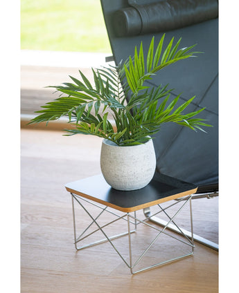 Ein künstlicher Palmensalon UV 60 cm steht in einem Topf auf einem kleinen modernen Beistelltisch mit schwarzer Platte und Metalldrahtbeinen, neben einem schwarzen Stuhl auf einem hellen Holzboden.