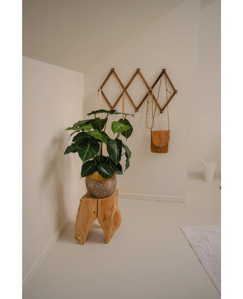 Een minimalistische kamerhoek wordt een groene oase met de Kunstplant Syngonium UV 58 cm op een krukje van natuurlijk hout, naast een bruine leren tas op een geometrisch wandrek en een witte kan, allemaal tegen gebroken witte muren en vloer.