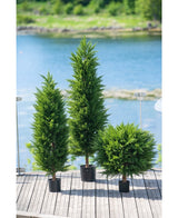 Drie Kunstplant Conifeer deluxe UV 120 cm bomen sieren een houten terras met glazen reling, met uitzicht op water en groen - ideaal voor elke kunstplant tuin dankzij hun UV-bescherming.
