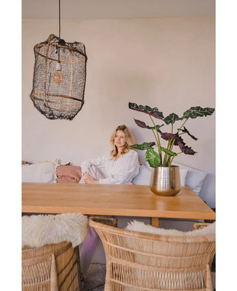 Een vrouw in een witte blouse zit aan een houten eettafel met rieten stoelen. De gezellige, zacht verlichte kamer heeft de Kunstplant Alocasia UV 67 cm en een hanglamp met textuur als decor.