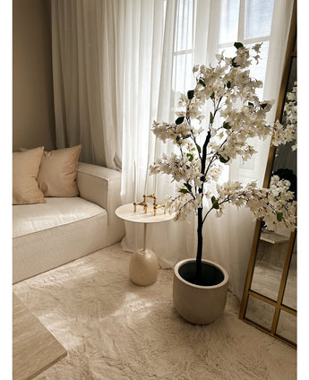 Een gezellige woonkamerhoek met een witte bank, beige kussens, een kleine ronde bijzettafel met gouden kandelaars, transparante gordijnen en de Kunstplant Bougainvillea 120 cm wit voor een elegante touch.