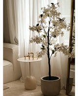 Een Kunstplant Bougainvillea van 120 cm groot staat bij een goudomrande spiegel naast een kleine ronde witte bijzettafel met gouden kandelaars, hoge witte kaarsen, transparante witte gordijnen en een crèmekleurige bank in een lichte kamer.