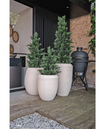 Drie hoge, witte plantenbakken met textuur en Kunstplant Conifeer UV 120cm kunstgroene planten staan op een houten terras naast een modern huis. Een zwarte buitengrill staat vlakbij, met een stenen muur en groen op de achtergrond.