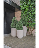 Drie hoge, lichtgekleurde plantenbakken met luxe buitenplanten zoals Kunstplant Conifeer deluxe UV 100 cm staan op een houten vlonder bij een met groene klimop begroeide muur, naast een zwarte deur en een gedeelte met gedeeltelijke grindgrond.