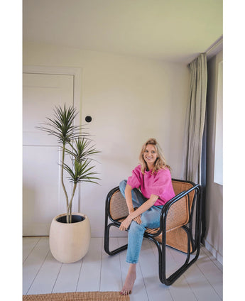 Eine Frau in einem knallrosa Top und blauen Jeans sitzt barfuß auf einem modernen schwarzen Korbstuhl neben der Kunstpflanze Dracaena Fragnans 150 cm in einem hellen, minimalistischen Raum mit weißen Wänden und Boden.