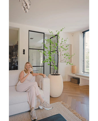 Eine Frau in heller Loungewear sitzt auf einem beigen Sofa und lächelt bei einer Tasse Kaffee in einem modernen Wohnzimmer mit Holzböden, raumhohen Fenstern und dem künstlichen Enkianthus 180 cm als große Topfpflanze.
