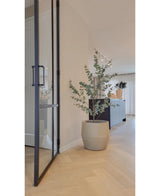 Een modern interieur met lichte houten vloeren, een grote glazen deur en de Kunstplant Eucalyptus 150 cm. Op de achtergrond staat een donkere kast terwijl zacht natuurlijk licht door de gordijnen naar binnen valt.