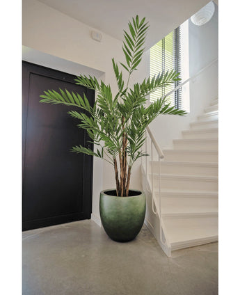 De Kunstplant Hawaii Parlour 180 cm, een hoge indoor kunstpalm met weelderige groene bladeren, staat op een grijze vloer in een moderne hal in de buurt van een witte trap en zwarte deur.