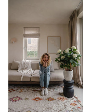 Een vrouw met krullend blond haar zit op een beige bank in een lichte woonkamer en laat haar kin op haar handen rusten. Naast haar staat de Kunstplant Hortensia UV 85 cm groot, verlicht door licht door een raam met beige gordijnen.