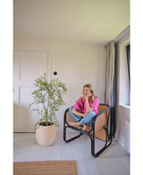 Eine Frau in rosa Hemd und Jeans sitzt barfuß auf einem geflochtenen Stuhl, lächelt und entspannt in einem hellen, minimalistischen Raum mit weißen Wänden und Böden. Neben ihr steht die künstliche Ruscus-Pflanze 120 cm.