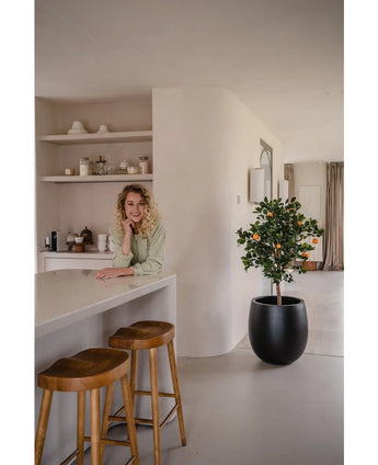 Een glimlachende vrouw met krullend blond haar leunt op het aanrecht in een moderne, lichte kamer met houten krukken, planken met serviesgoed en een Kunstplant Sinaasappelboom 120 cm.