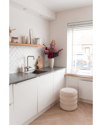 Een moderne, minimale keuken met witte kastjes, een zwart aanrechtblad en een Kunstbloem Hortensia 55 cm donkerroze in een vaas bij het aanrecht, plus houten planken met decor en een ronde kruk met kussens bij een raam met jaloezieën.