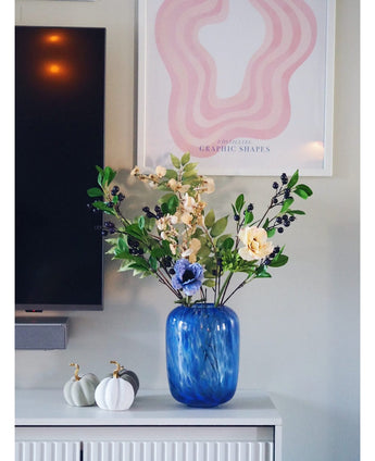 Een blauwe glazen vaas met een Kunsttak Berry 76 cm paars kunstbloem staat op een witte kast. Ernaast staan twee witte pompoenen met gouden stelen. Aan de muur erachter hangen een tv en een abstracte roze kunstprint.