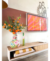 Auf einem Holzregal steht eine farbenfrohe Vase mit der Art Branch Mandarin 93 cm orange, daneben ein gerahmtes Kunstwerk mit rosa Streifen, auf dem „Kissje op je Hart“ steht. Außerdem stehen auf dem Regal eine grüne Kerze und gefaltete Decken.