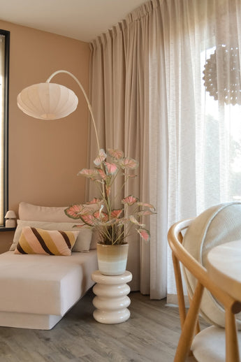 Eine gemütliche Sitzecke mit einer beigen Chaiselongue, einem gestreiften Kissen, dem künstlichen Caladium 90 cm in Rosa, das für einen Hauch von Grün sorgt, einer modernen Lampe, Lichtvorhängen und einem Holzboden. Durch die transparenten Vorhänge fällt sanftes natürliches Licht ein.