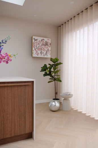 Een moderne, minimale hoek met de Kunstplant Ficus Tabaksplant Deluxe 155 cm, een klein wit krukje, ronde zilveren plantenbak, beige gordijnen, houten kast, muurkunst en vaas met bloemen op een lichthouten vloer.